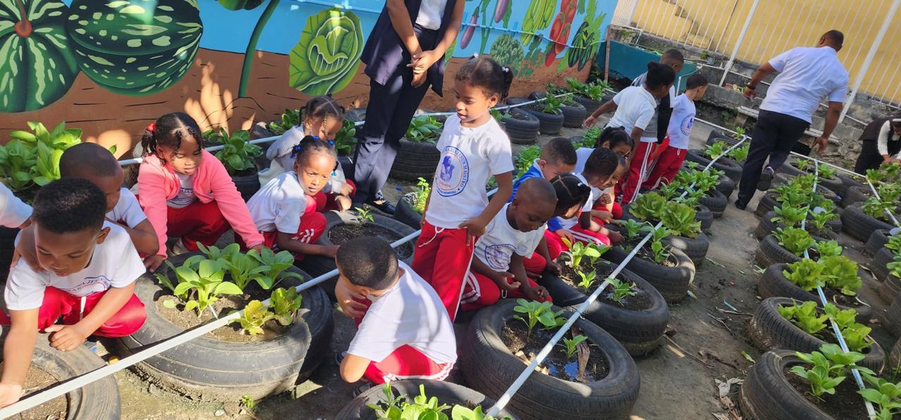 Young children kneeling along rows of recycled tires planted with seedlings, in a school courtyard beneath a painted mural of fruit and vegetables.